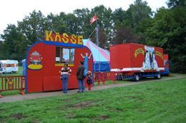 Roter Zirkuswagen mit Kasse, Besuchergruppe und Zelt im Hintergrund auf grünem Rasen.
