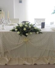 Tavolo rotondo decorato con tovaglia bianca, fiori e candelabri. Sfondo con piante floreali.