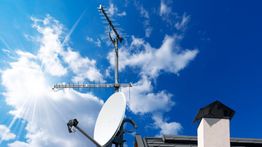 Antena parabólica en un techo bajo un cielo azul con nubes y sol brillante.