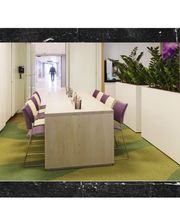 Moderner Büroraum mit Tisch, lila Stühlen und Pflanzen; Person im Hintergrund.