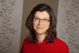 Eine Frau mit Brille und rotem Hemd lächelt vor dezent gemustertem Hintergrund in die Kamera.