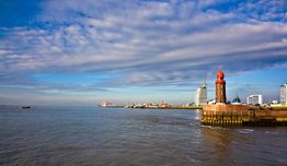 Leuchtturm am Wasser mit Stadtansicht und bewölktem Himmel im Hintergrund.