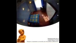 Una stanza con letto matrimoniale, tenda blu e pavimento rosso; logo Comune di Lucca.