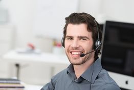 Mann mit Headset lächelt im Büro, Computer im Hintergrund.