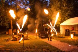 Feuershow im Freien mit Jongleur, Flammen und Zelt bei Nacht, Bäume im Hintergrund.