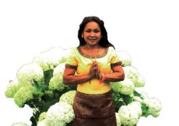 Frau in traditioneller Kleidung vor weißen Blumen, Hände in Grußhaltung zusammengelegt.