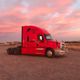 Red semi-truck parked on gravel under a pink and orange sunset sky.