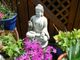 Buddha-Statue in einem Garten, umgeben von bunten Blumen und Pflanzentöpfen vor einem Holzzaun.