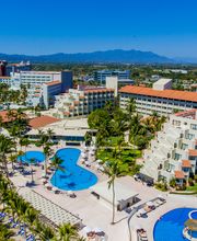 Vista aérea de un complejo hotelero junto a la playa, con piscinas y palmeras bajo cielo despejado.