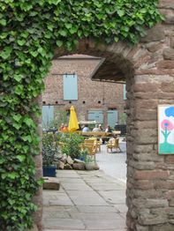 Blick durch ein steinernes Tor in einen begrünten Innenhof mit Besuchern und gelbem Sonnenschirm.