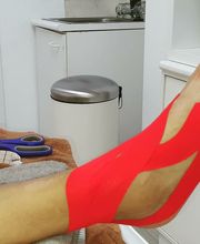 Pie con cinta kinesiológica roja en un consultorio. Tijeras y material médico al fondo.