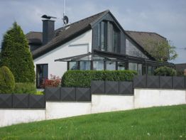 Modernes Einfamilienhaus mit Wintergarten, umgeben von Garten und Hecke, an einem sonnigen Tag.
