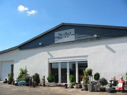 Außenansicht eines Ladens mit Pflanzen und Gartendekorationen auf dem Gehweg bei sonnigem Wetter.