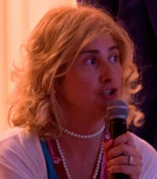 Persona con microfono, capelli biondi ricci e collana di perle, parla animatamente.