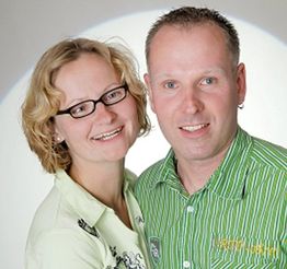 Ein lächelndes Paar posiert vor einem neutralen Hintergrund. Beide tragen Hemden in hellen Farben.