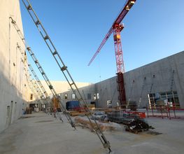 Baustelle mit Betonmauern und rotem Kran bei klarem Himmel. Gerüste und Baumaterialien sichtbar.