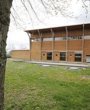 Holzgebäude neben grünem Rasen und Baum im Vordergrund. Überdachter Bereich rechts.