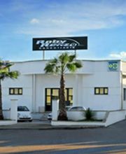 Edificio bianco con insegna sul tetto e due palme davanti, cielo sereno e soleggiato.