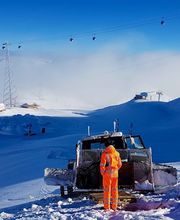 Person in orange neben einem Schneepflug auf schneebedecktem Berg unter blauem Himmel.