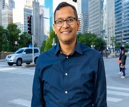 Smiling man in glasses and blue shirt standing on a city street with skyscrapers in the background.