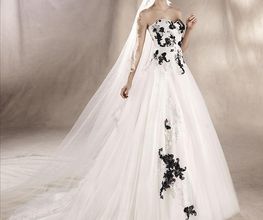 Braut in weißem Kleid mit schwarzen Blumenmustern und langem Schleier vor neutralem Hintergrund.