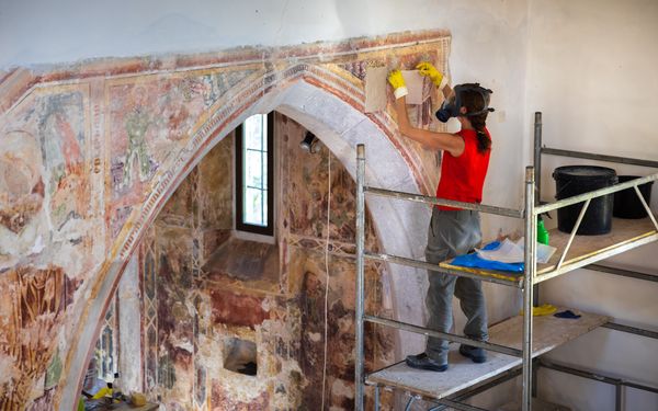 Restaurierungsprozess an einer Kirchenbogenwand durch eine erfahrene Restauratorin, die eine schützende Atemmaske trägt, während sie die ursprünglichen Freskenfarben mit Hilfe einiger chemischer Behandlungen hervorbringt.