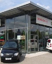 Autohändler mit Glasfassade, zwei Autos davor, Himmel im Hintergrund.