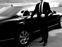 Man in a suit standing by a luxury car, holding the door handle, by a lakeside road.