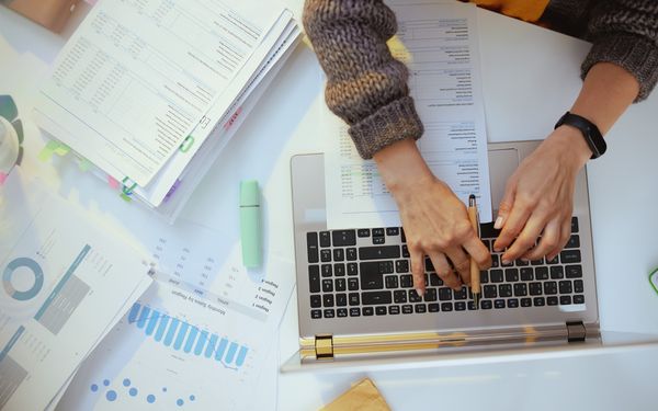Weibliche Hände tippen auf der Tastatur eines Laptops steuerlich bezogene Dinge ein. Neben dem Laptop liegen zwei Stapel an gekennzeichneten Dokumenten.