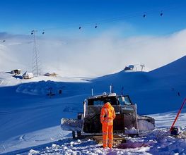 Person in orange neben einem Schneepflug auf schneebedecktem Berg unter blauem Himmel.