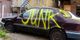 Old car with "JUNK" spray-painted in yellow on the side, parked beside urban buildings.