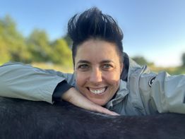 Person lächelt in die Kamera und lehnt sich auf ein schwarzes Tier, draußen bei klarem Himmel.