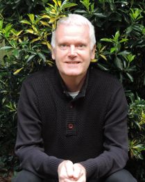 Een man met wit haar, zittend voor groene struiken, draagt een donkere trui en glimlacht in de camera.