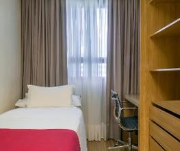 Quarto simples com cama de solteiro, escrivaninha e estante de madeira ao lado da janela.