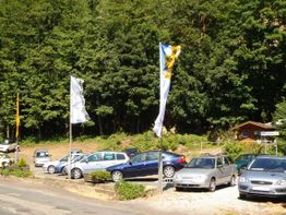 Autos parken vor einem Wald, daneben wehen Fahnen. Eine kleine Holzhütte steht im Hintergrund.