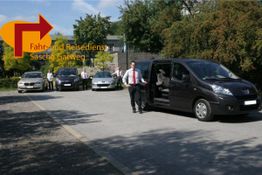 Ein Mann steigt aus einem schwarzen Van aus, vier Autos im Hintergrund, Logo oben links.