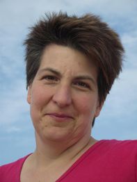 Porträt einer Frau mit kurzen Haaren und rotem Oberteil, lächelt vor blauem Himmel.