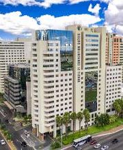 Conjunto de prédios modernos em uma cidade, com céu azul e algumas nuvens. Ruas movimentadas abaixo.