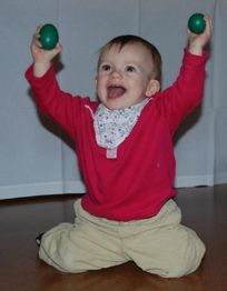 Ein lachendes Kleinkind hält zwei grüne Eier in den Händen und trägt eine rote Jacke.