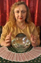 Frau mit Kristallkugel und Tarotkarten auf einem Tisch, vor rotem Vorhang sitzend.