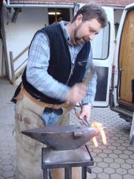 Mann schmiedet glühendes Eisen auf einem Amboss neben einem weißen Lieferwagen.