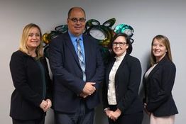 Four people in business attire stand smiling in front of a decorative wall piece.