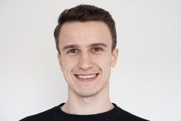 Ein lächelnder Mann vor weißem Hintergrund, trägt ein dunkles Oberteil.