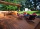 Wooden deck with pergola, red cushioned chairs, tables, and bench, surrounded by greenery.