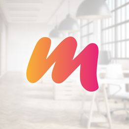 Buntes "M"-Logo vor einem unscharfen Bürohintergrund mit Schreibtischen und Hängelampen.