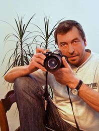 Mann mit Kamera vor Pflanzen, in lässiger Pose, trägt helles Oberteil und Jeans.