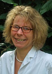 Frau mit Brille und lockigem Haar lächelt, trägt weiße Bluse vor grünem Hintergrund.