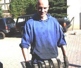 Eine Person hält ein großes Zahnrad, im Freien, mit Autos im Hintergrund.