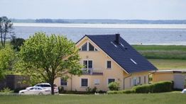 Gelbes Haus am See mit grüner Wiese und Baum im Vordergrund an einem sonnigen Tag.