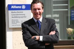 Ein Mann im Anzug steht vor einem Allianz-Schild und lächelt in die Kamera.